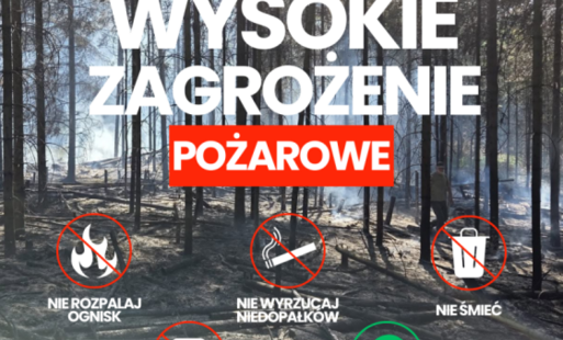 Na zdjęciu znajduje się plakat ostrzegawczy dotyczący wysokiego zagrożenia pożarowego w lesie. W tle znajduje się las, w którym widać ślady po pożarze lub akcji gaśniczej – ziemia jest zwęglona, a między drzewami unosi się dym. U góry umieszczono czerwony